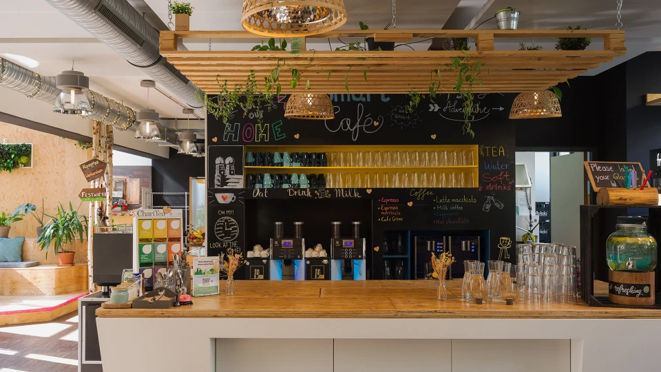 Innenansicht eines gemütlichen Cafés mit einer Theke, darüber eine Tafel mit handgeschriebenen Menüs, umgeben von Holzregalen und hängenden Pflanzen.