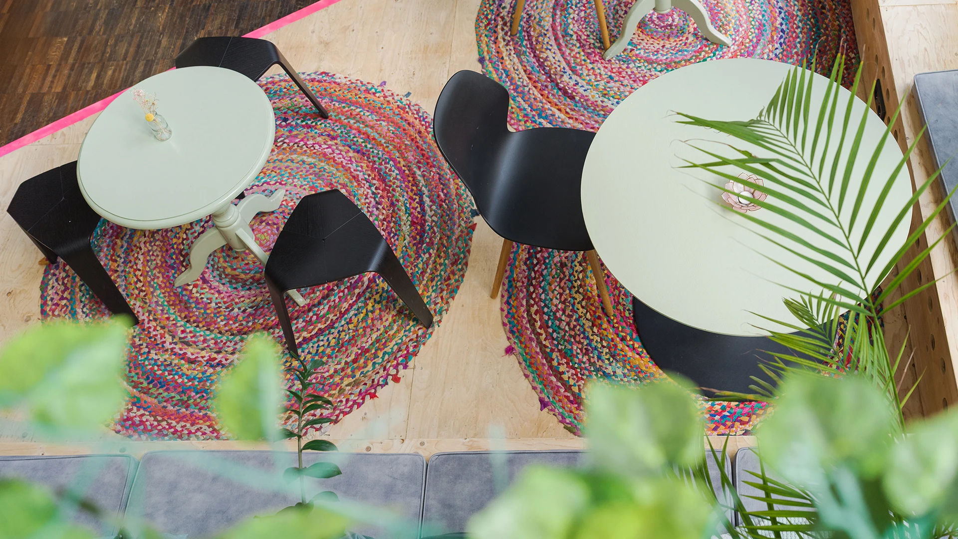 Draufsicht auf ein Café mit bunten, runden Teppichen und kleinen Tischen, umgeben von grünen Pflanzen