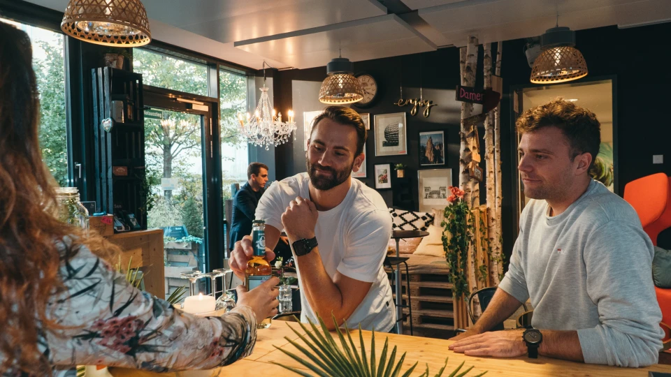 Zwei Männer stehen an einer Bar und unterhalten sich, im Hintergrund ist eine gemütliche Bar-Atmosphäre mit Dekoration und Hängelampen.