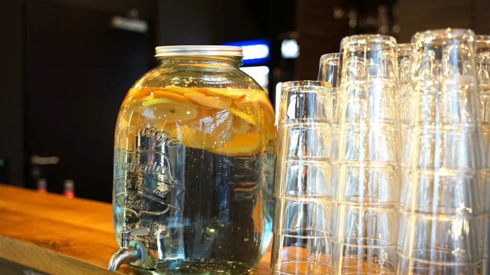 Ein großer Glasbehälter mit Wasser und Orangenscheiben steht auf einer Theke, daneben sind saubere Gläser aufgestapelt.