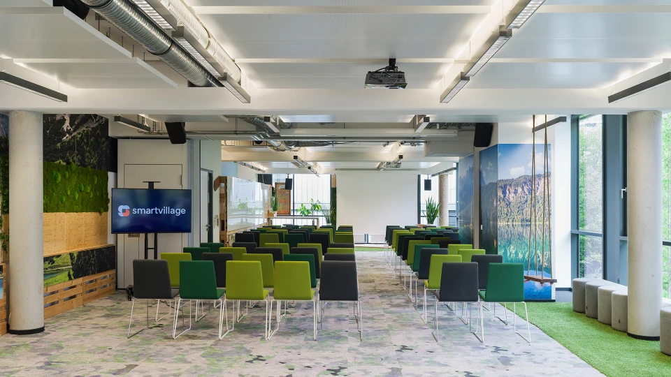 Moderne Büroumgebung mit grünen und grauen Stühlen, angeordnet für ein Meeting. Im Hintergrund große Bilder von Naturmotiven und das Logo 'smartvillage'.