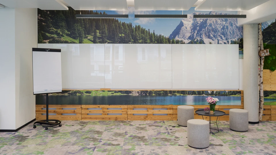 Modernes Büro mit Alpenpanorama an den Wänden, ausgestattet mit einem Flipchart, runden Sitzhockern und einem Blumenarrangement.