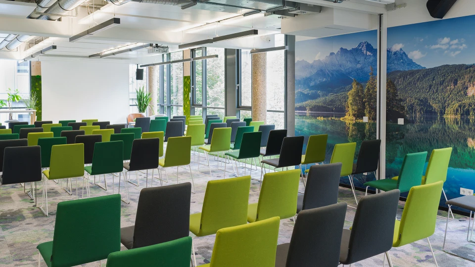 Moderne Konferenzraumgestaltung mit grünen und grauen Stühlen, gerichtet auf eine Vortragsfläche, umgeben von großen Fenstern und Naturfotografien an den Wänden.