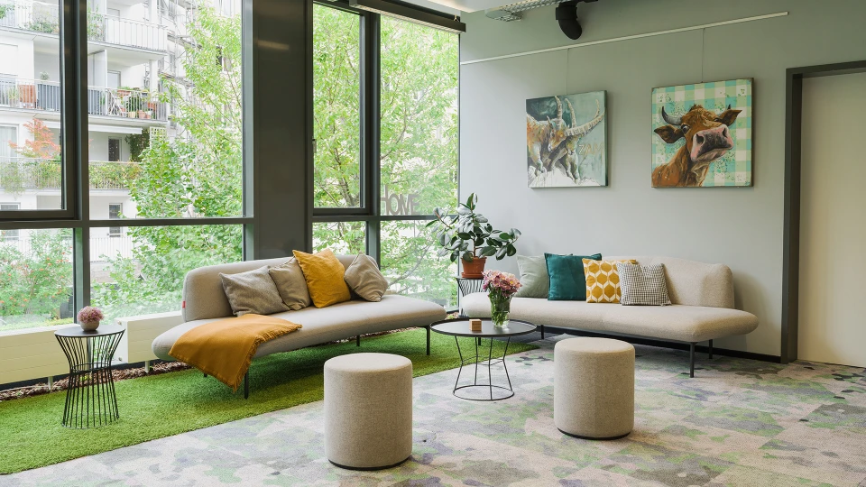 Modernes Wohnzimmer mit zwei Sofas, bunten Kissen, einem runden Couchtisch, zwei Hockern und Kunstwerken an der Wand neben großen Fenstern mit Blick auf grüne Bäume.