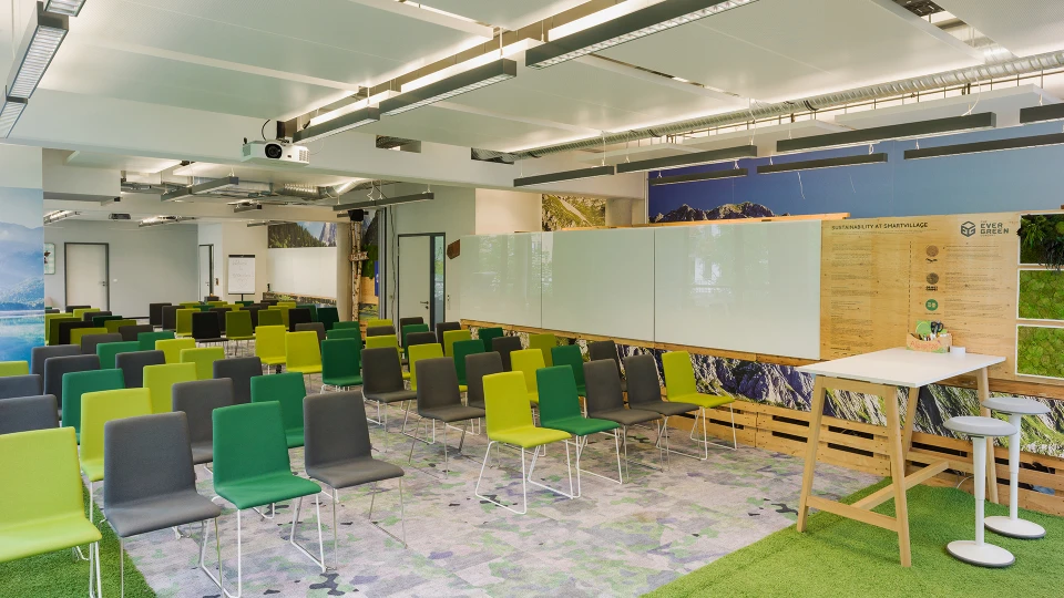Modernes Büro-Seminarraum mit grünen und grauen Stühlen, einem großen Holztisch und informativen Wandtafeln.