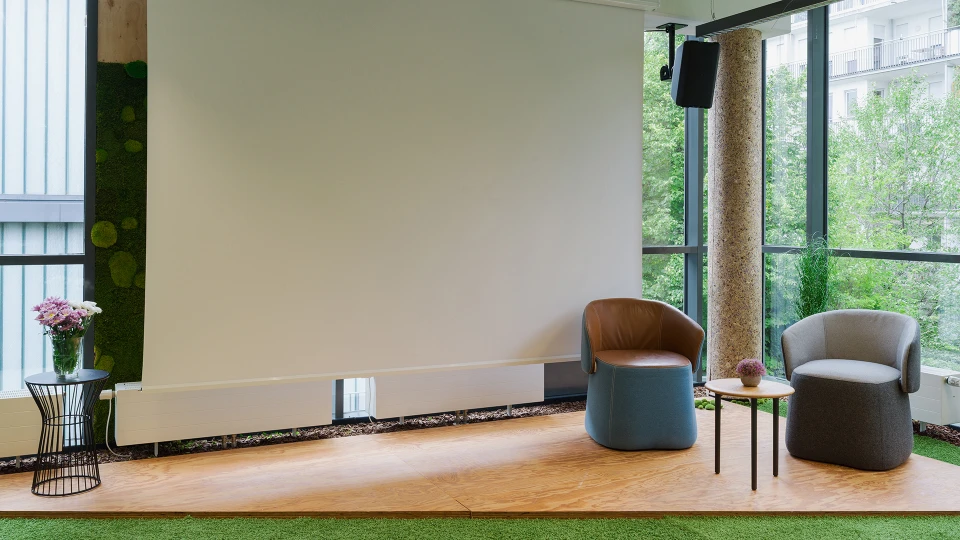 Modernes Lounge-Design mit zwei Sesseln und einem Tisch vor einer großen weißen Leinwand, umgeben von großen Fenstern mit Blick auf grüne Bäume.