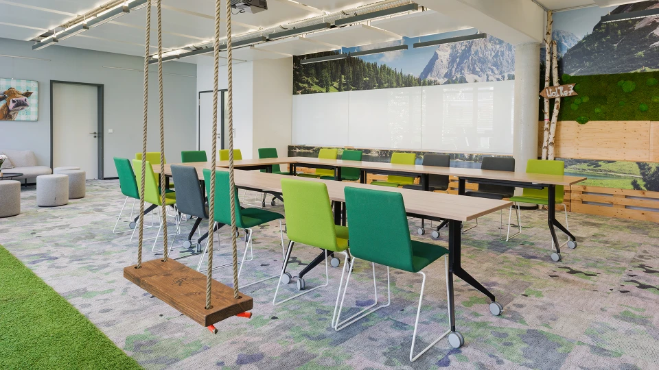Modernes Büro mit einem langen Tisch und grünen sowie grauen Stühlen, Schaukeln an der Decke und Bergpanorama-Wandbild