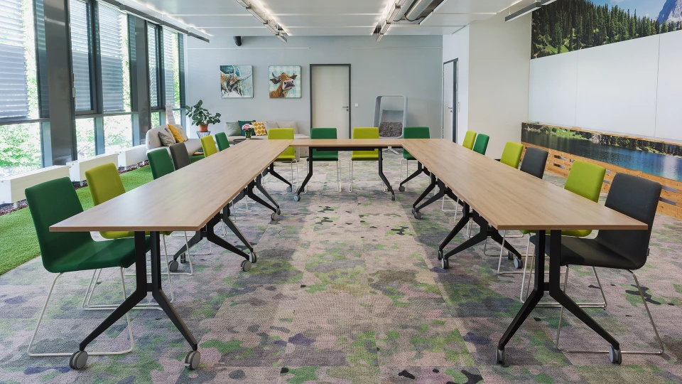 Modernes Bürokonferenzzimmer mit einem langen Tisch, grünen und gelben Stühlen, umgeben von Kunstwerken und einer Glaswand mit Blick auf Grünflächen.