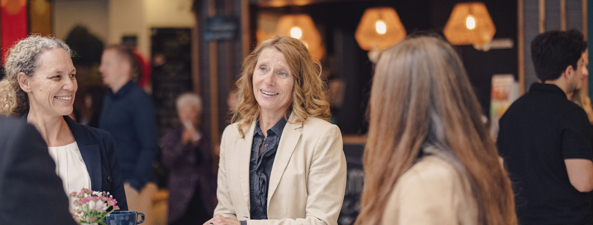 Drei Frauen unterhalten sich in einem belebten Raum mit stilvoller Beleuchtung, zwei lächeln, während die dritte ihnen zuhört.