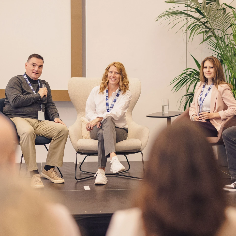 Drei Personen auf einer Podiumsdiskussion in einem Konferenzraum, sitzend vor einem Publikum. Zwei Frauen und ein Mann, lächelnd, tragen Namensschilder und lässige Geschäftskleidung.