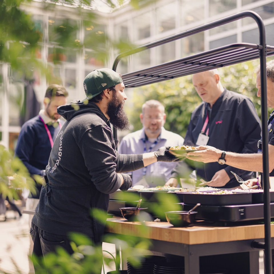 Mann mit Bart und Kappe serviert Essen an Gäste an einem Buffet im Freien in einem glasüberdachten Bereich