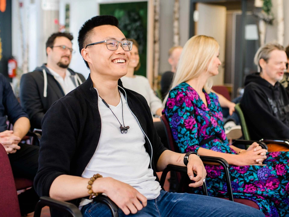 Lachender junger Mann mit Brille sitzt in einer Gruppe von Zuschauern bei einer Veranstaltung.