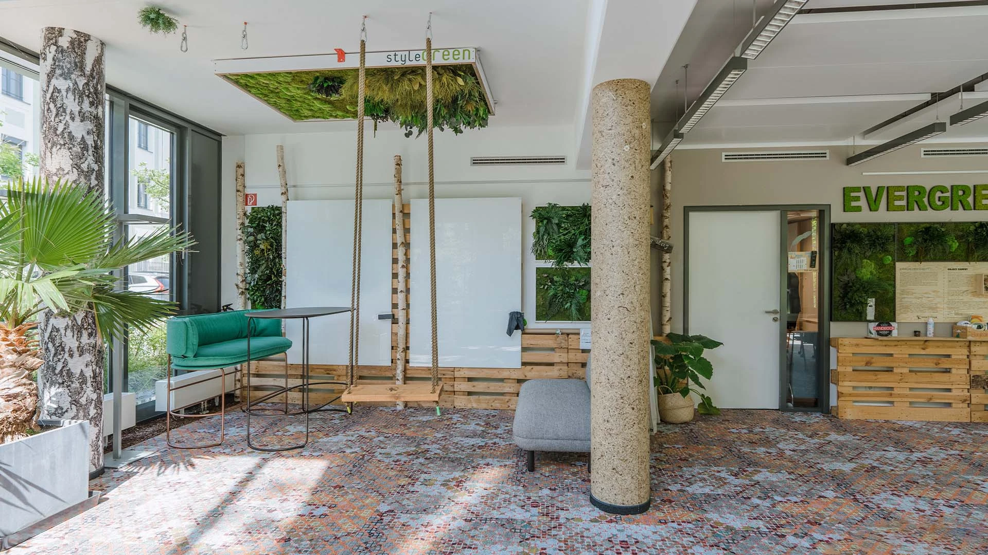 Modernes Bürofoyer mit üppiger Begrünung, einschließlich Hängepflanzen und Wandbegrünung. Ausstattung mit gemütlichem grünen Sofa, grauem Hocker und einer Schaukel. Raumgestaltung betont Nachhaltigkeit.