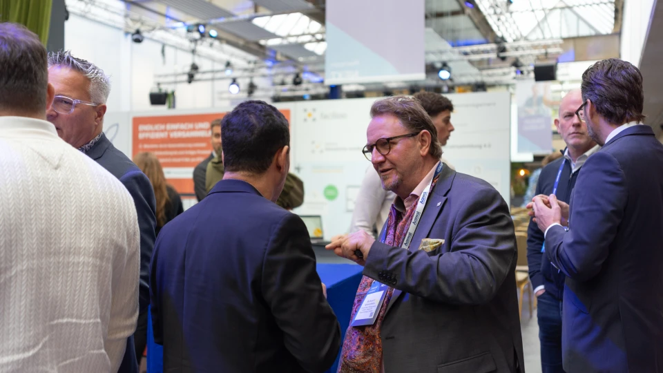 Mehrere Geschäftsleute unterhalten sich aktiv auf einer Messe mit blauem Teppich und Informationsständen im Hintergrund.