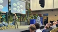 Mann präsentiert auf einer Konferenz vor Publikum, steht auf einer Bühne neben einem Bildschirm mit Grafiken. Moderne, helle Innenraumgestaltung.