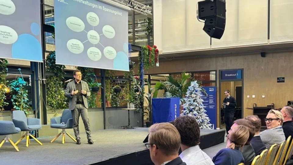 Mann präsentiert auf einer Konferenz vor Publikum, steht auf einer Bühne neben einem Bildschirm mit Grafiken. Moderne, helle Innenraumgestaltung.