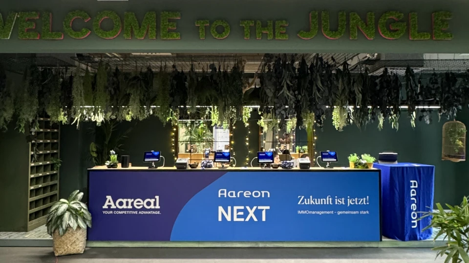 Empfangsbereich mit 'Welcome to the Jungle' Schild, verziert mit grünen Pflanzen, hinter einem Tresen mit 'Aareon NEXT' Banner.