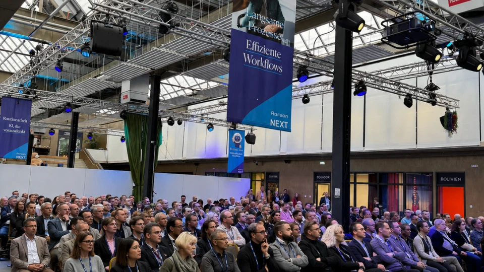 Publikum sitzt in einer modernen Halle mit Glasdach bei einer Konferenz, umgeben von Bannern und technischer Beleuchtung.