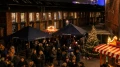 Abendlicher Weihnachtsmarkt mit beleuchteten Ständen und einer Menschenmenge, die sich um Tische unter Sonnenschirmen versammelt, in einem urbanen, industriell wirkenden Umfeld.