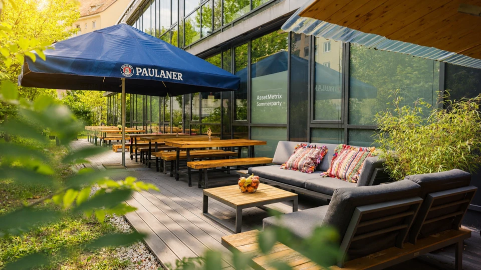 Außenterrasse eines Cafés mit einem großen blauen Paulaner-Sonnenschirm, hölzernen Bierzeltgarnituren und einer gemütlichen Sitzecke mit Sofa und Kissen.