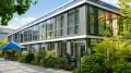 Modernes zweistöckiges Glasgebäude umgeben von grünen Bäumen und Sträuchern mit einem blauen Sonnenschirm im Vordergrund.
