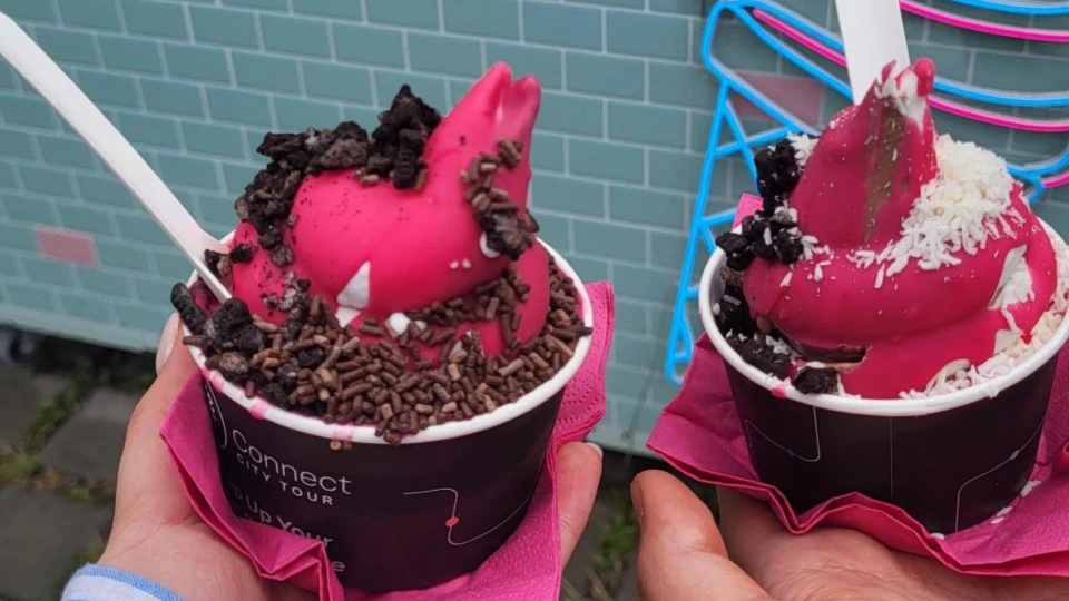 Zwei Hände halten je einen Becher mit pinkem Eis, dekoriert mit Schokostreuseln und Kokosraspeln, vor einer türkisfarbenen Fliesenwand.