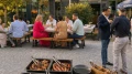 Menschen essen und unterhalten sich an Tischen im Freien bei einem Grillbuffet mit verschiedenen Fleischsorten auf einem Stadtplatz.