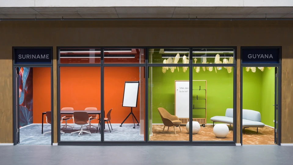Zwei moderne Bürokonferenzräume mit Glasfronten, beschriftet mit 'SURINAME' in Rot und 'GUYANA' in Grün, ausgestattet mit Stühlen, Tischen und Dekorationselementen.