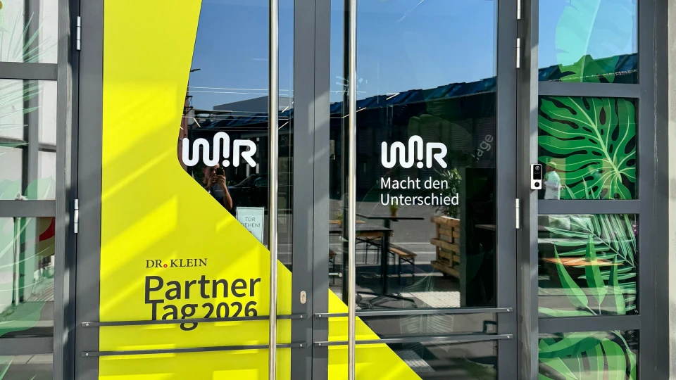Glastüren eines Gebäudes mit gelben Bannern, die 'WIR Dr. Klein Partner Tag 2026' und 'WIR Macht den Unterschied' ankündigen.