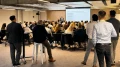 Geschäftsmann präsentiert vor einer Gruppe sitzender und stehender Zuhörer in einem modernen Konferenzraum