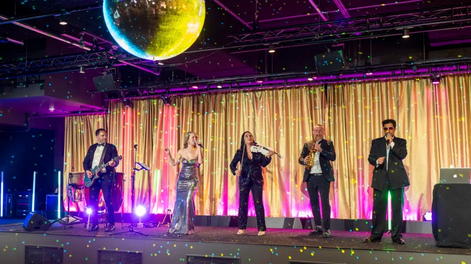 Band mit sechs Mitgliedern, darunter Sänger und Musiker, auf einer Bühne bei einem Event, beleuchtet von Bühnenlichtern und einer Discokugel