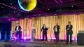 Band mit sechs Mitgliedern, darunter Sänger und Musiker, auf einer Bühne bei einem Event, beleuchtet von Bühnenlichtern und einer Discokugel