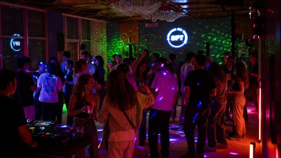 Menschen tanzen und unterhalten sich in einem lebhaften Nachtclub mit bunten Lichtern und einem DJ-Pult.