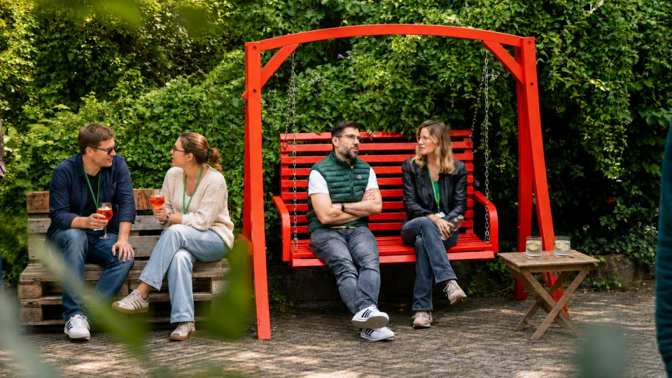 Zwei Paare, die sich auf einer roten Schaukelbank unterhalten, umgeben von grüner Hecke im Hintergund