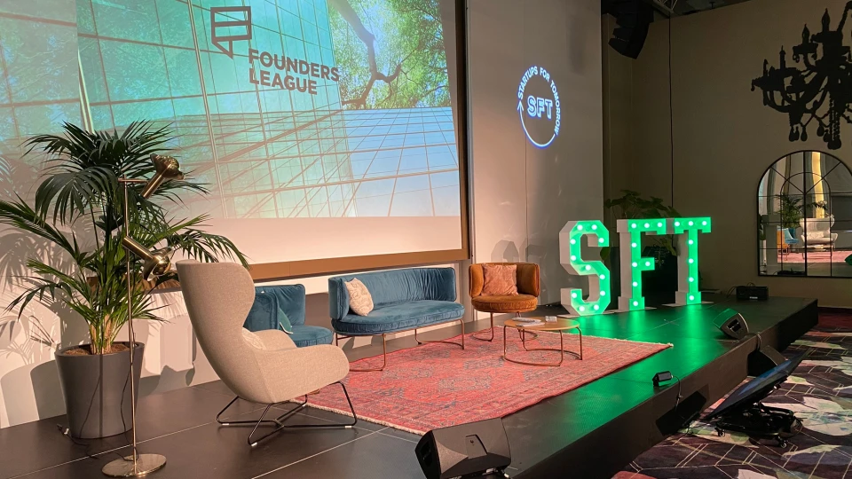 Eine Konferenzbühne mit SFT-Leuchtschild, vier Sesseln, darunter ein Teppich, zwei Pflanzen und eine Präsentationsleinwand mit der Aufschrift 'Founders League'.