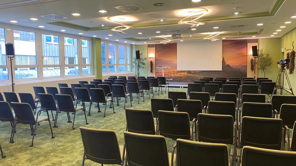 Leerer Konferenzraum mit Reihen von schwarzen Stühlen, die auf eine große Leinwand an der Vorderseite gerichtet sind. Der Raum hat grünen Teppich und Tageslicht durch Fenster.