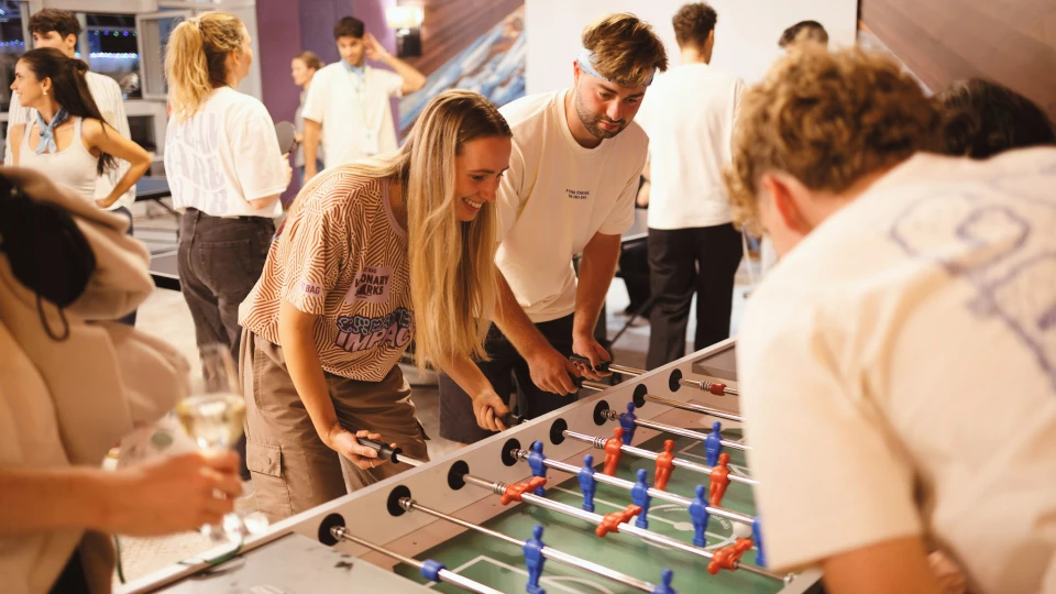 Eine Gruppe junger Erwachsener spielt Tischfußball in einem belebten Raum, zwei Männer und eine Frau sind im Vordergrund aktiv am Spiel beteiligt.