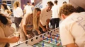 Eine Gruppe junger Erwachsener spielt Tischfußball in einem belebten Raum, zwei Männer und eine Frau sind im Vordergrund aktiv am Spiel beteiligt.