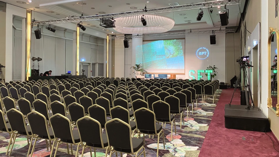 Großer Konferenzsaal mit vielen leeren Stühlen, Bühne mit Großbildschirm und grünen dekorativen Elementen