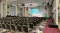 Großer Konferenzsaal mit vielen leeren Stühlen, Bühne mit Großbildschirm und grünen dekorativen Elementen