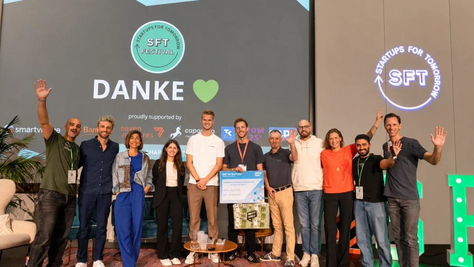 Gruppe von elf Personen auf einer Bühne bei einem Startup-Festival, sie lächeln und einige winken, im Hintergrund steht 'DANKE' und Logos von Sponsoren