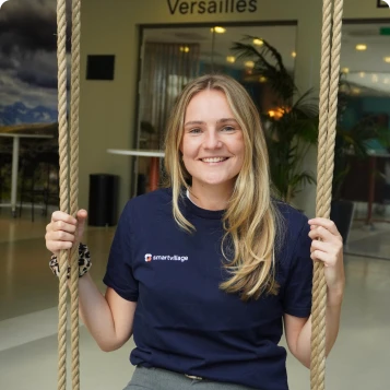 Junge Frau mit langen blonden Haaren lächelt in die Kamera und hält sich an den Seilen einer Schaukel fest, im Hintergrund ein Schild mit der Aufschrift 'Versailles'.