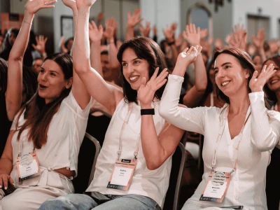 Drei Frauen in weißer Kleidung, die bei einer Konferenz sitzen und begeistert die Hände heben