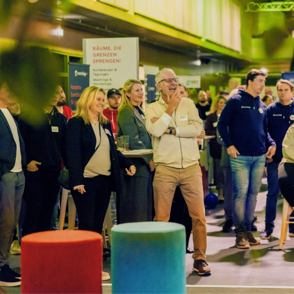 Menge von Menschen, darunter ein älterer Mann und eine Frau, die lächelnd bei einer Veranstaltung stehen, mit einem Schild im Hintergrund, das 'RÄUME, DIE GRENZEN SPRENGEN' liest
