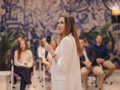 Frau in weißem Anzug hält eine Rede mit Mikrofon vor sitzendem Publikum in einem Raum mit blauem Wandgemälde